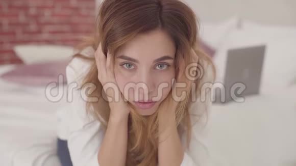 一个精疲力竭的白人女孩坐在床上用手抱着她的头的肖像美丽的女人看着视频的预览图