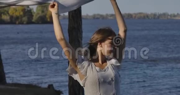 一位身穿浅色连衣裙的白种人美丽女子站在岸上的肖像闭着眼睛举着围巾视频的预览图