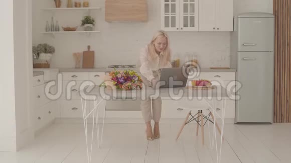 快乐的白人女士金发站在现代厨房看着笔记本电脑做胜利的手势成人视频的预览图