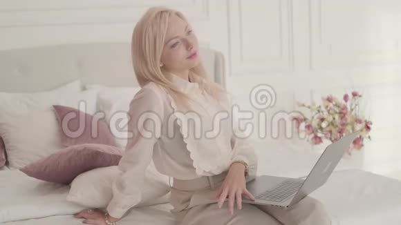 笑容满面的金发灰眼白种人女商人坐在床上拿着笔记本电脑思考年轻漂亮的女士视频的预览图