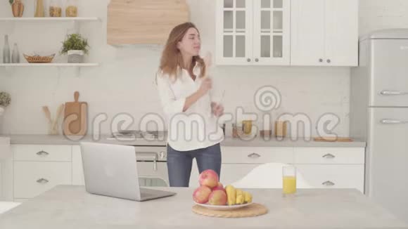 快乐的白人女孩穿着休闲服装在轻盈的现代厨房里用木制勺子跳舞快乐美丽的女人视频的预览图