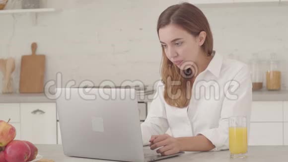 年轻疲惫的白种人女商人坐在厨房里手提电脑叹气穿白色衬衫的年轻漂亮女孩视频的预览图
