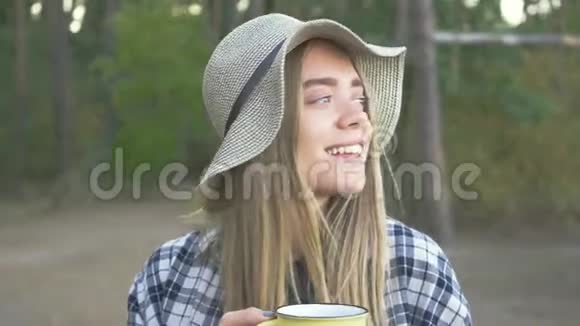 年轻的白种人女孩穿着格子衬衫坐在森林里拿着一个黄色的杯子微笑着户外休闲视频的预览图