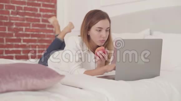 年轻的白人女人躺在床上在笔记本电脑上打字吃红苹果在家学习的漂亮女学生视频的预览图