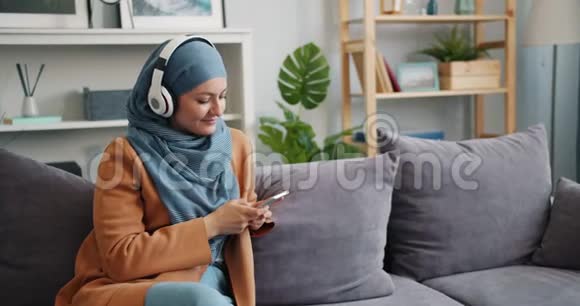 戴着头巾的穆斯林女孩在公寓里用智能手机听音乐视频的预览图