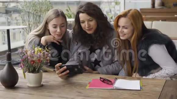 三个白种人的女朋友围着毯子坐在咖啡馆里讨论手机上的照片青年妇女视频的预览图