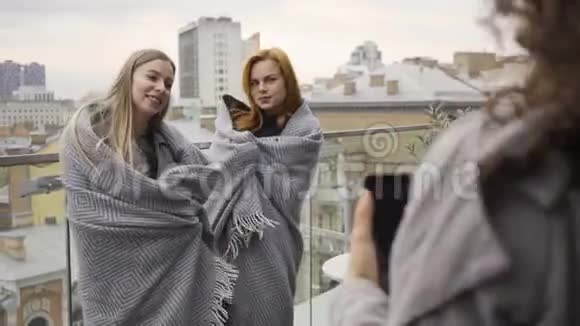 两个白种人的女朋友在阳台上站着被毯子覆盖着为她拍照的女人视频的预览图