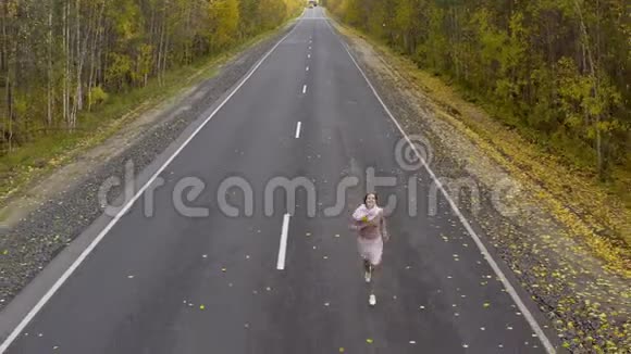 穿着粉红色衣服的女人在秋天森林中的沥青路上奔跑和旋转顶部视图视频的预览图
