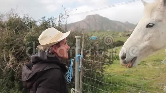 白马在绿色的草地上从他的女牛仔那里得到一根胡萝卜戴着白色的牛仔帽用嘴直接喂给他视频的预览图