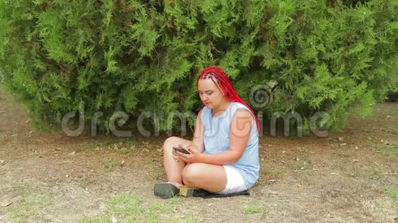 一个年轻的女人坐在草地上的公园里看着她的手机视频的预览图