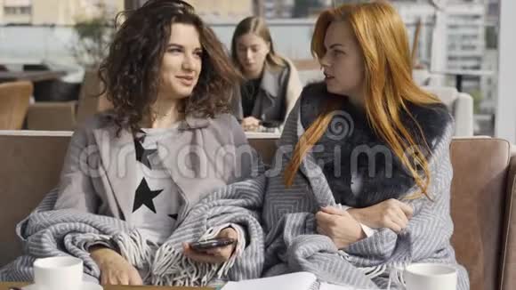 两个漂亮的白种人女朋友围着毯子坐在咖啡馆里讨论手机上的照片青年妇女视频的预览图