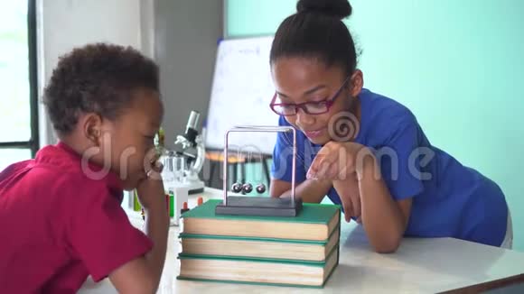 两个非裔美国人混合孩子用放大镜和拉牛顿的摇篮球摆动在科学教室视频的预览图