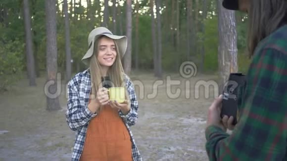 年轻的白种人妇女戴着草帽在森林的背景上摆着黄色的杯子男朋友在拍照片视频的预览图