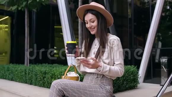 慢动作可爱浪漫的年轻女士戴着时髦的帽子和时髦的衣服坐在混凝土栏杆上使用视频的预览图
