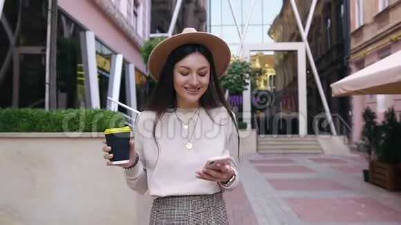 漂亮的快乐满意的女人戴着时髦的帽子走在办公楼附近街边的咖啡馆里一边用手机视频的预览图