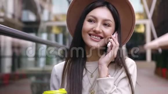 美丽的微笑快乐的女人戴着时尚的帽子走在玻璃墙附近享受着她的电话交谈视频的预览图