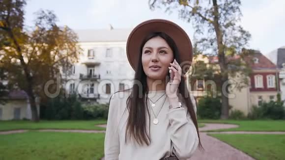 穿着时髦帽子的可爱漂亮女人在豪华的草坪上漫步时一边打电话一边说话视频的预览图