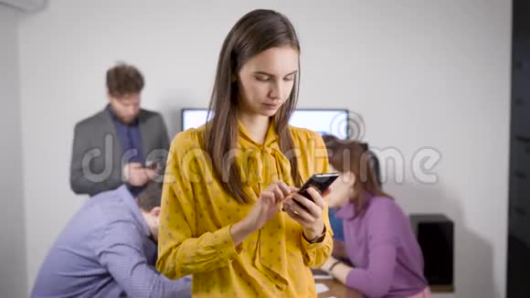 年轻女子站在办公室的智能手机上打字同事们坐在后台视频的预览图