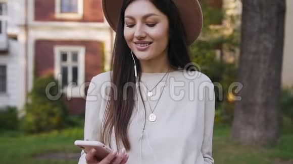 美丽的微笑现代女性戴着时尚的帽子走在美丽的富人家附近用耳机听音乐视频的预览图