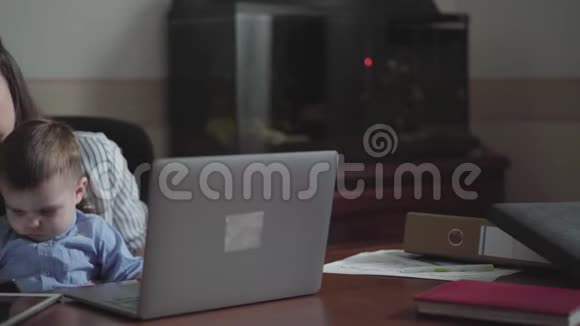 小儿子坐在妈妈桌旁的大腿上用妈妈的手摸手机忙碌的年轻母亲在家工作这就是视频的预览图