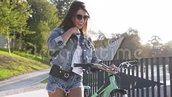 美丽的微笑女孩戴着时尚的太阳镜穿着时髦的衣服在阳光明媚的日子里骑着自行车靠近栅栏视频的预览图