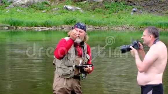 渔夫和摄影师站在河里一位留着长发和圣诞老人胡须的老人选择了一个捕蝇鱼饵视频的预览图