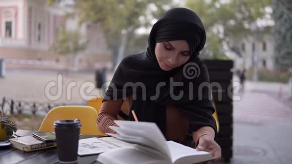 一个迷人的穆斯林女孩在一本笔记本上写字她坐在咖啡馆里拿着一本书在户外咖啡馆学习休息视频的预览图