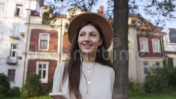浪漫的迷人的苗条的戴着可爱帽子的年轻女子走在漂亮的房子附近听着耳机里的音乐视频的预览图