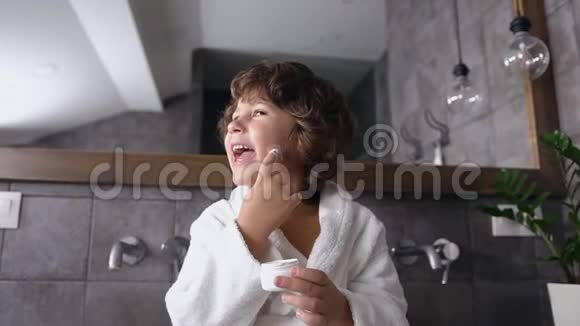 可爱的小男孩卷发穿着白色长袍坐在浴室镜子附近玩护肤霜视频的预览图