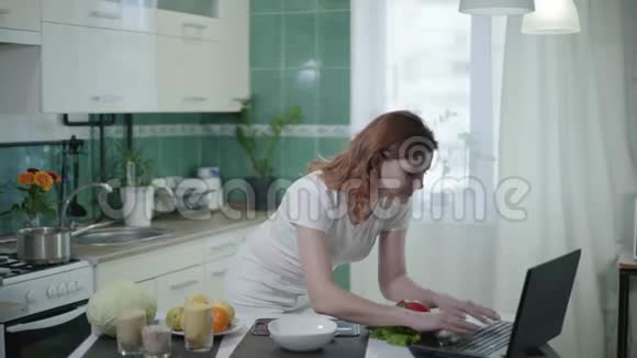 家庭主妇在厨房煮健康食品时使用笔记本电脑女孩用勺子测量麦粥并观看视频的预览图