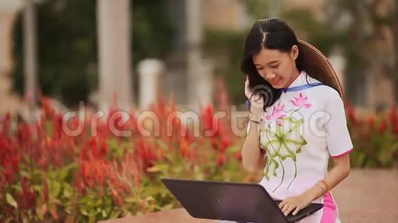 漂亮的亚洲女孩坐在公园里手里拿着一台笔记本电脑穿着国服戴着一顶帽子戴在诺拉的背上视频的预览图