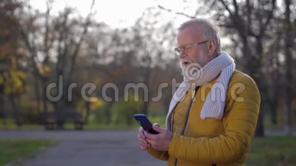 一位留着灰胡子手里拿着手机的现代祖父在秋天散步时通过视频通话进行交流视频的预览图