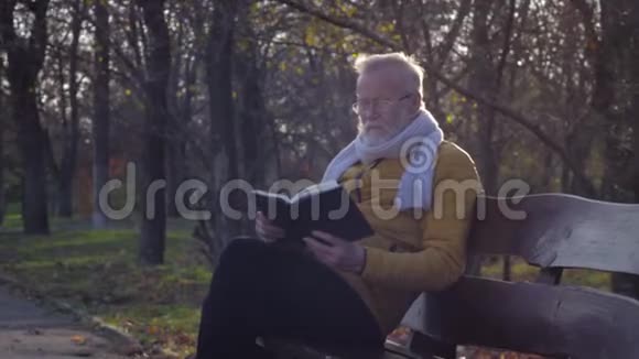 退休休闲中年老人戴着眼镜在秋天的公园里坐在长椅上看书视频的预览图
