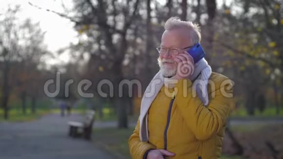 秋天公园散步时留着灰色胡子的快乐老人在手机上说话的肖像视频的预览图