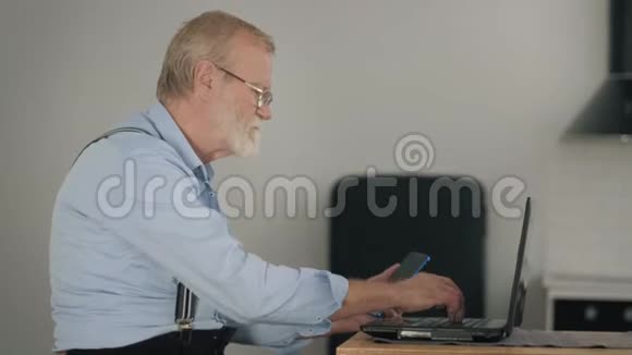 现代的老年人忙碌的老人戴着眼镜在网上支付账单坐在办公桌前用笔记本电脑和手机视频的预览图
