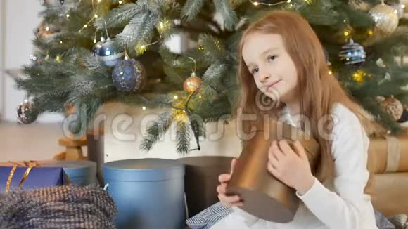 微笑着可爱的金发女孩拥抱着一个金色的礼盒她坐在圣诞树旁快乐的童年和圣诞节视频的预览图