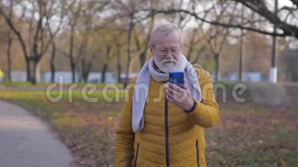 老人用手机打视频电话男的留着灰胡子走在公园里展现了大自然的美丽视频的预览图
