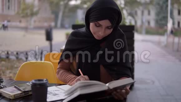 一个迷人的穆斯林女孩在一本笔记本上写字她坐在咖啡馆里拿着一本书在户外咖啡馆学习休息视频的预览图