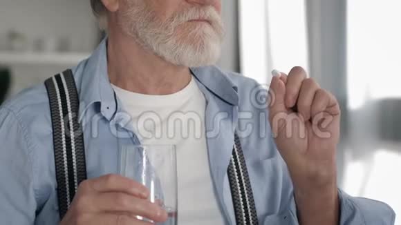长着灰胡子的老人在白色的房间里吃药丸喝杯子里的水来保持健康动作缓慢视频的预览图