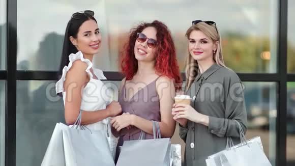 可爱的三个时尚女孩带着购物袋微笑着看着镜头摆在玻璃窗上视频的预览图