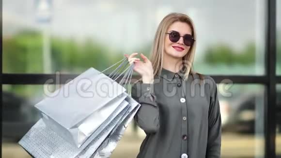 一位身着时尚太阳镜摆着购物袋的金发女郎随意微笑视频的预览图