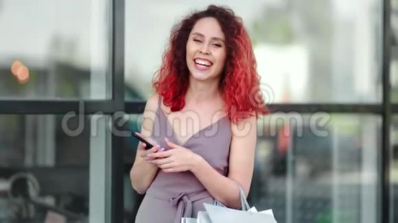 肖像微笑美丽的红头发年轻的购物狂带着智能手机看相机视频的预览图