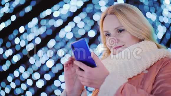 年轻女子在圣诞装饰的夜晚灯光背景下使用智能手机视频的预览图