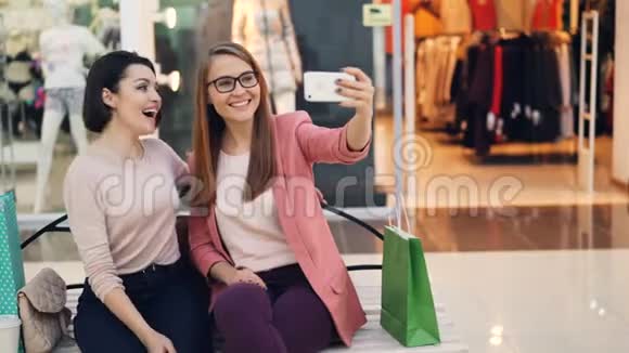 快乐的女性朋友用智能手机自拍坐在商场的长凳上戴着眼镜摆姿势视频的预览图