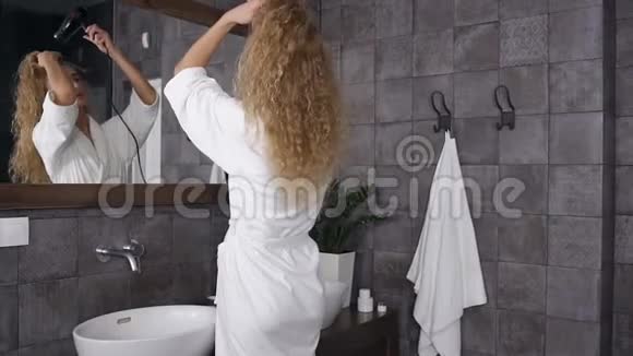 漂亮的金发女郎穿着白色长袍站在浴室的镜子旁边用吹风机吹干她卷曲的头发视频的预览图