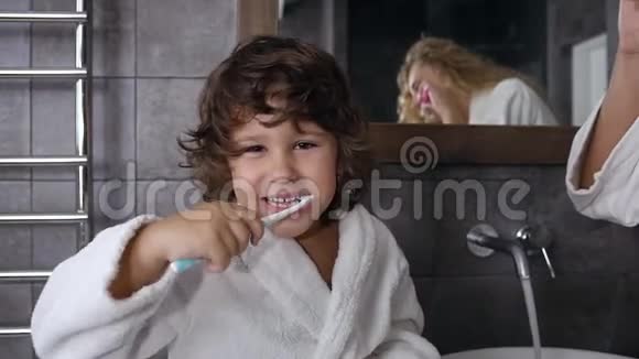 微笑的小男孩穿着长袍一头深色卷发在浴室镜子附近刷牙视频的预览图