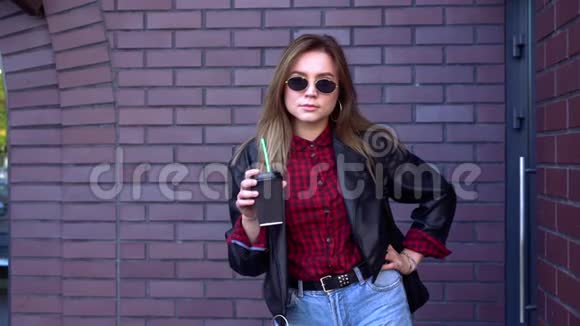秋天年轻漂亮的女学生站在砖墙附近戴着太阳镜手里拿着咖啡笑着视频的预览图