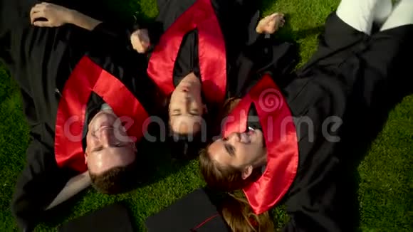 三个快乐微笑的学生穿着黑色斗篷躺在草地上享受她的自由的肖像青年视频的预览图