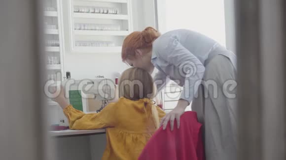 小女孩学习如何在靠近窗户的桌子旁缝制衣服而她的红发母亲视频的预览图