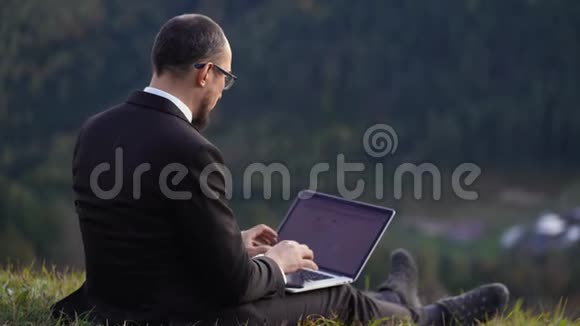 一个戴眼镜穿西装的男人坐在山上膝上放着笔记本电脑视频的预览图